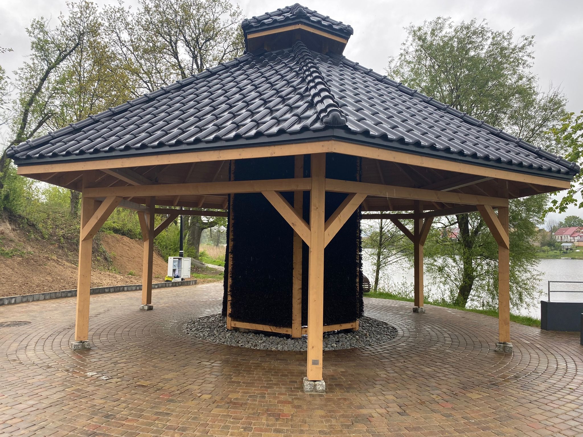 Otwarta altana z ciemnym dachem krytym dachówką, drewnianą konstrukcją i centralnie umieszczoną ścianą z tarniny, otoczona brukowanym placem i zielenią.