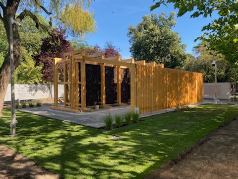 Drewniana konstrukcja solankowa w parku, z elementami zadaszenia i ścianką, otoczona trawnikiem i ścieżką. Jasne drewno, błękitne niebo w tle.