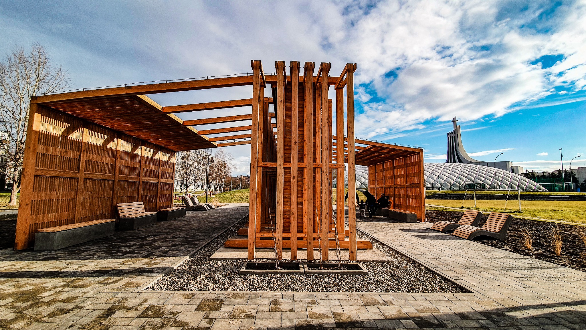 Nowoczesna, drewniana konstrukcja z zadaszeniem w parku miejskim. Elementy z drewna tworzą ażurową ścianę. W tle widoczny charakterystyczny budynek.