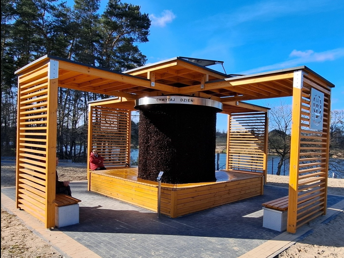 Drewniana konstrukcja solankowa z napisem 'Chwytaj Dzień' w parku, z widocznymi elementami drewnianymi i osobami siedzącymi na ławkach. Jasne, słoneczne oświetlenie.