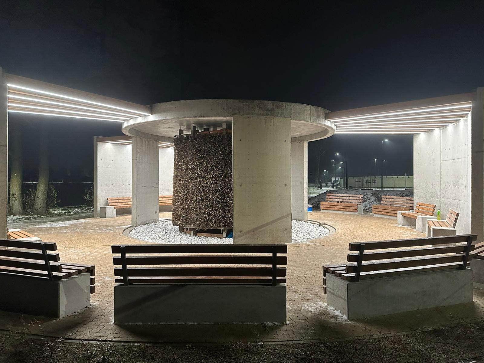 Oświetlona, betonowa altana z tężnią solankową w centrum, otoczona ławkami. Nocne ujęcie z niskiego kąta, podkreślające nowoczesny design i oświetlenie.