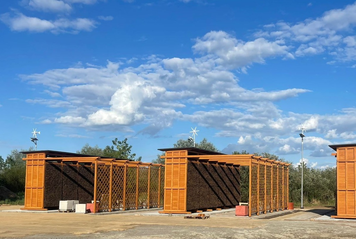 Kompleks tężni solankowych z drewnianą konstrukcją i miniaturowymi turbinami wiatrowymi na tle błękitnego nieba z chmurami. Widok z perspektywy lekko z dołu.