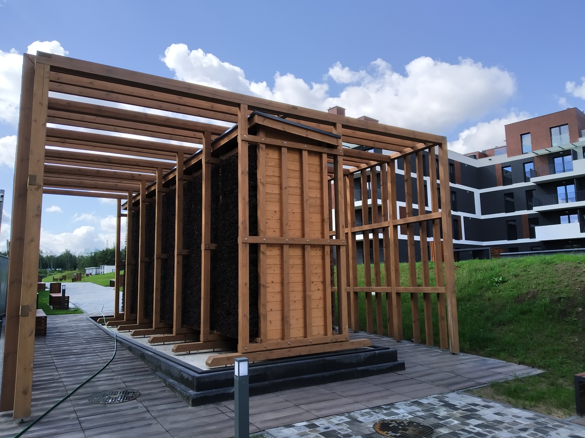 Drewniana konstrukcja solankowa z elementami z drewna i tarniny, na tle nowoczesnego budynku i błękitnego nieba z chmurami. Ujęcie z poziomu chodnika.