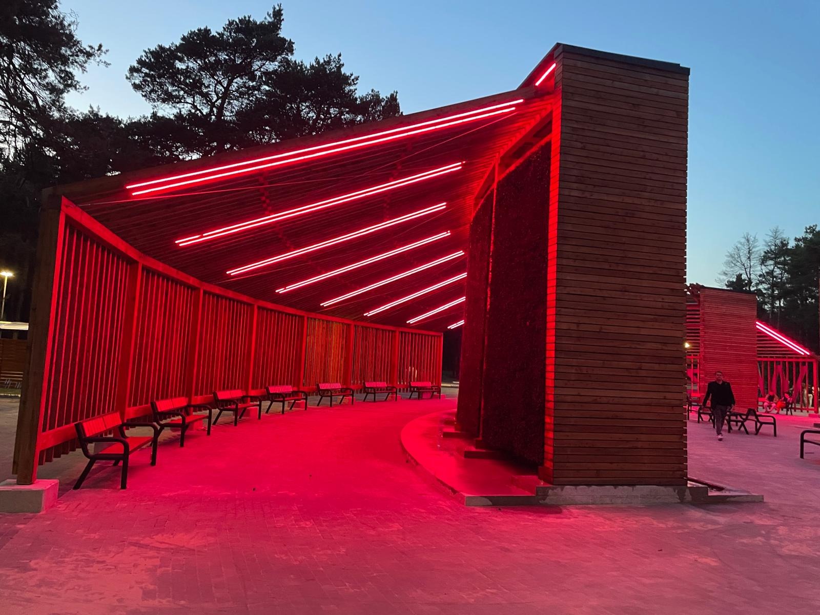Nowoczesna altana z drewna z czerwonym oświetleniem LED, ławkami i elementami solankowymi, otoczona drzewami. Wieczorny krajobraz.