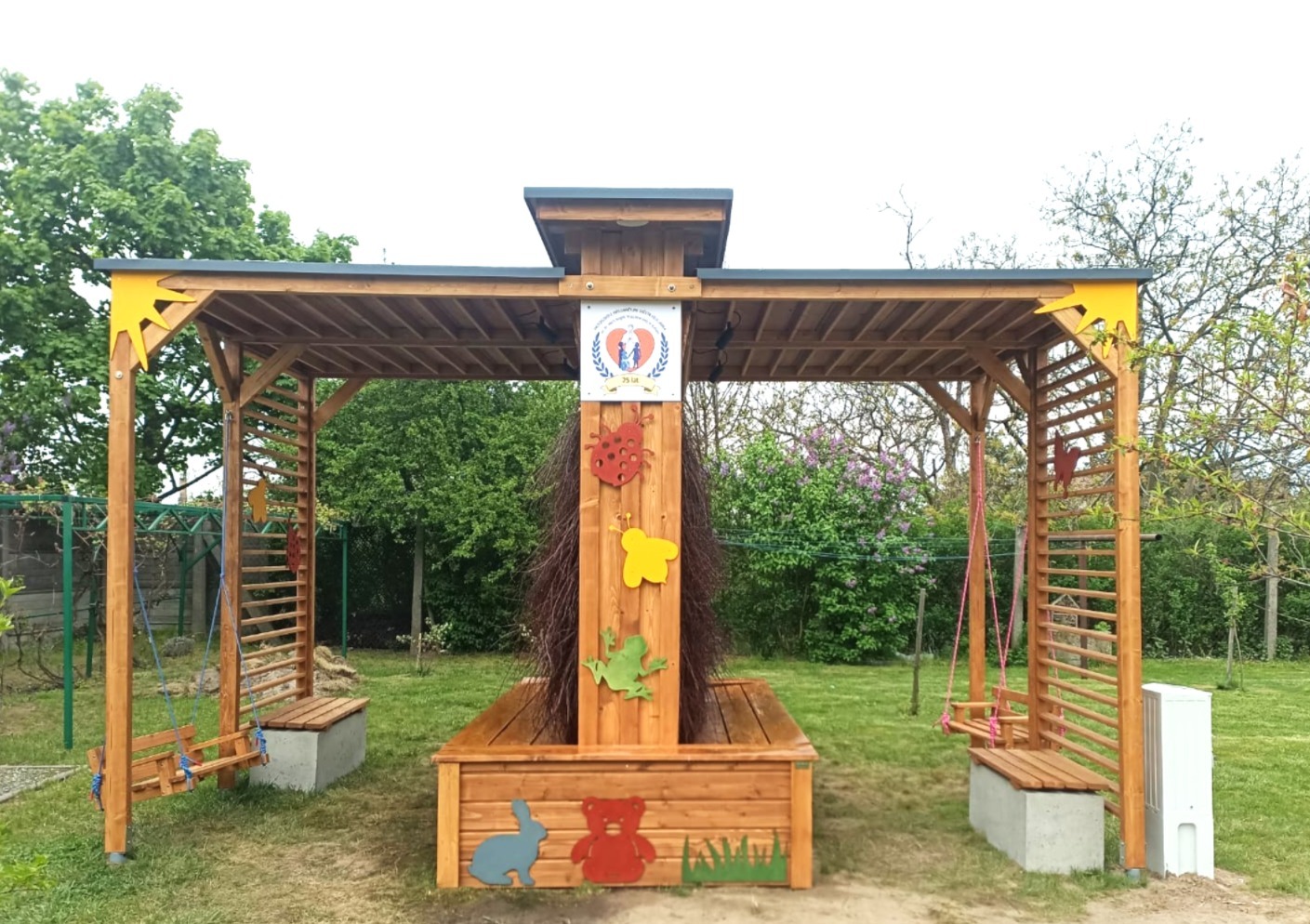 Drewniana konstrukcja z huśtawkami i dekoracjami dla dzieci w Koszalinie. Elementy pomalowane na żółto. Widoczne motywy zwierząt i roślin.