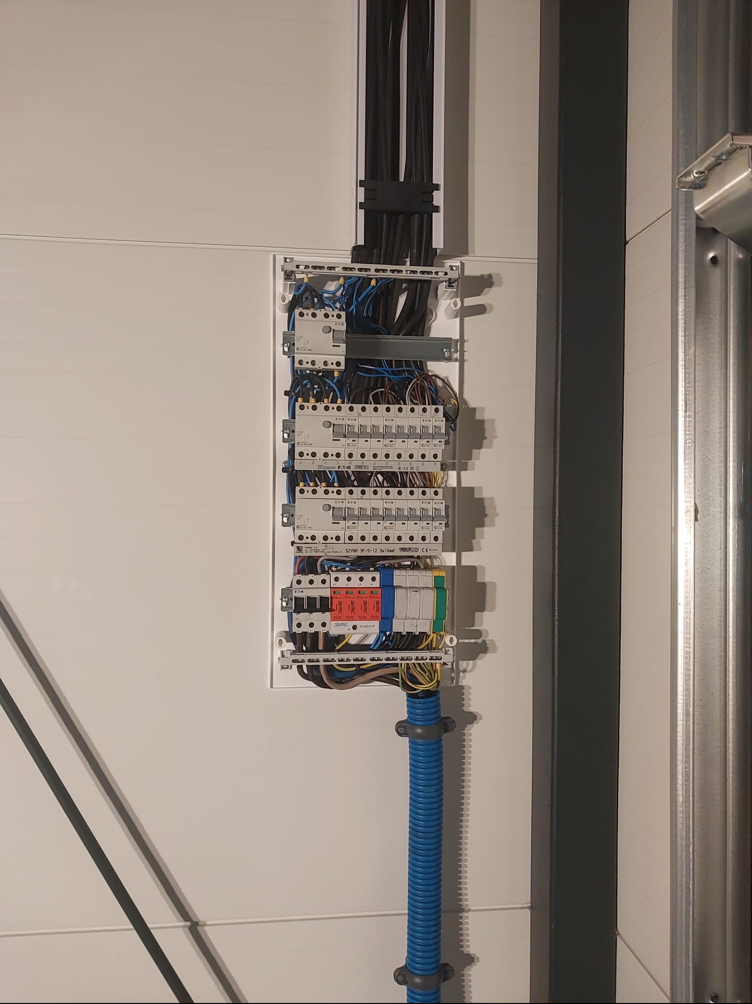 Rozdzielnia elektryczna z zabezpieczeniami, widoczne przewody w peszlu. Biała ściana w tle. Elementy instalacji zamontowane na szynie DIN.