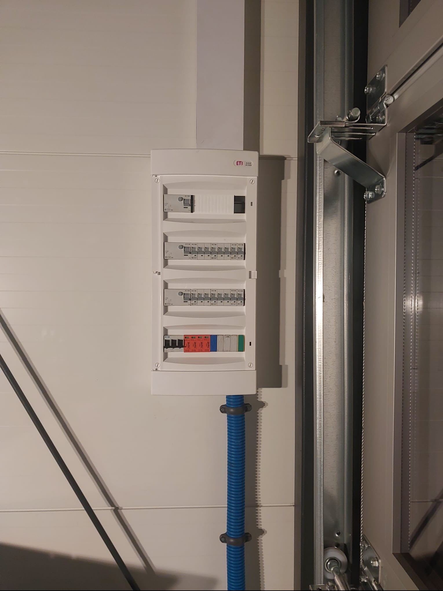 Zamknięta, biała skrzynka rozdzielcza ETI zamontowana na białej ścianie z niebieską rurą karbowaną, widoczna część metalowej konstrukcji bramy garażowej.