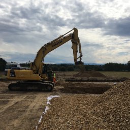 Marcin Baranowicz - Żółta koparka Komatsu 210 w trakcie prac ziemnych, widoczne wykopane rowy i hałda żwiru, operator w kabinie, pochmurne niebo w tle.