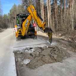 Marcin Baranowicz - Żółta koparko-ładowarka JCB z pneumatycznym młotem rozbija betonową nawierzchnię drogi. W tle las i błękitne niebo.