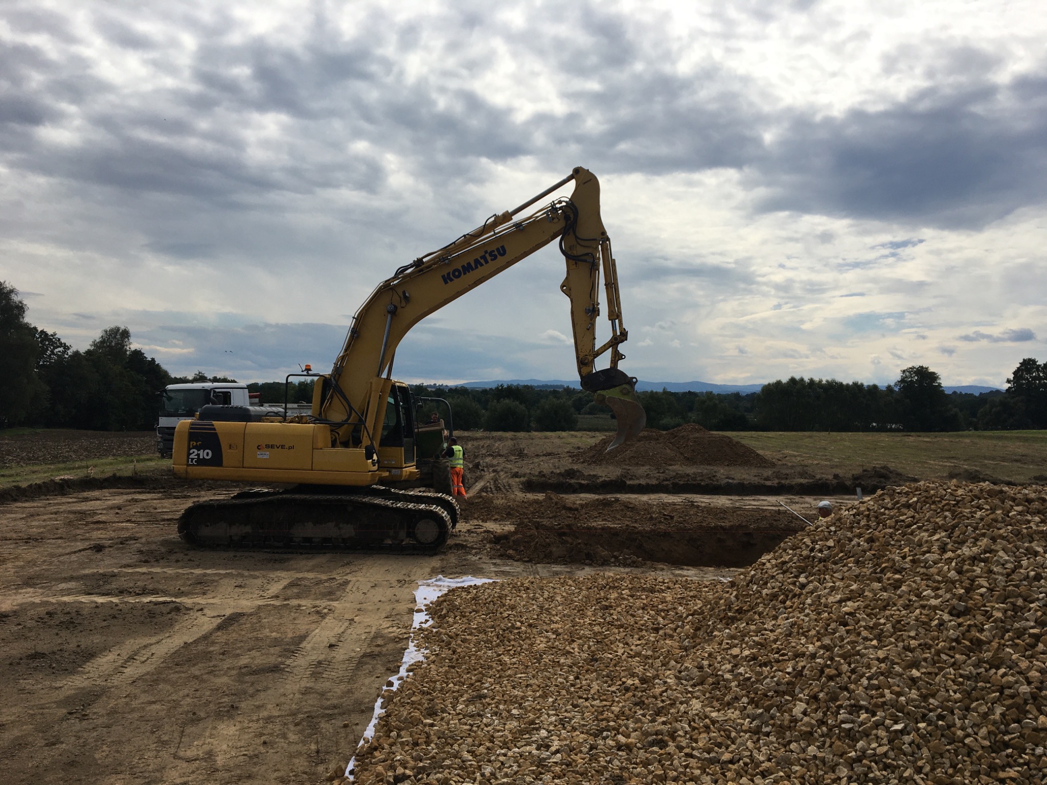 Żółta koparka Komatsu 210 w trakcie prac ziemnych, widoczne wykopane rowy i hałda żwiru, operator w kabinie, pochmurne niebo w tle.