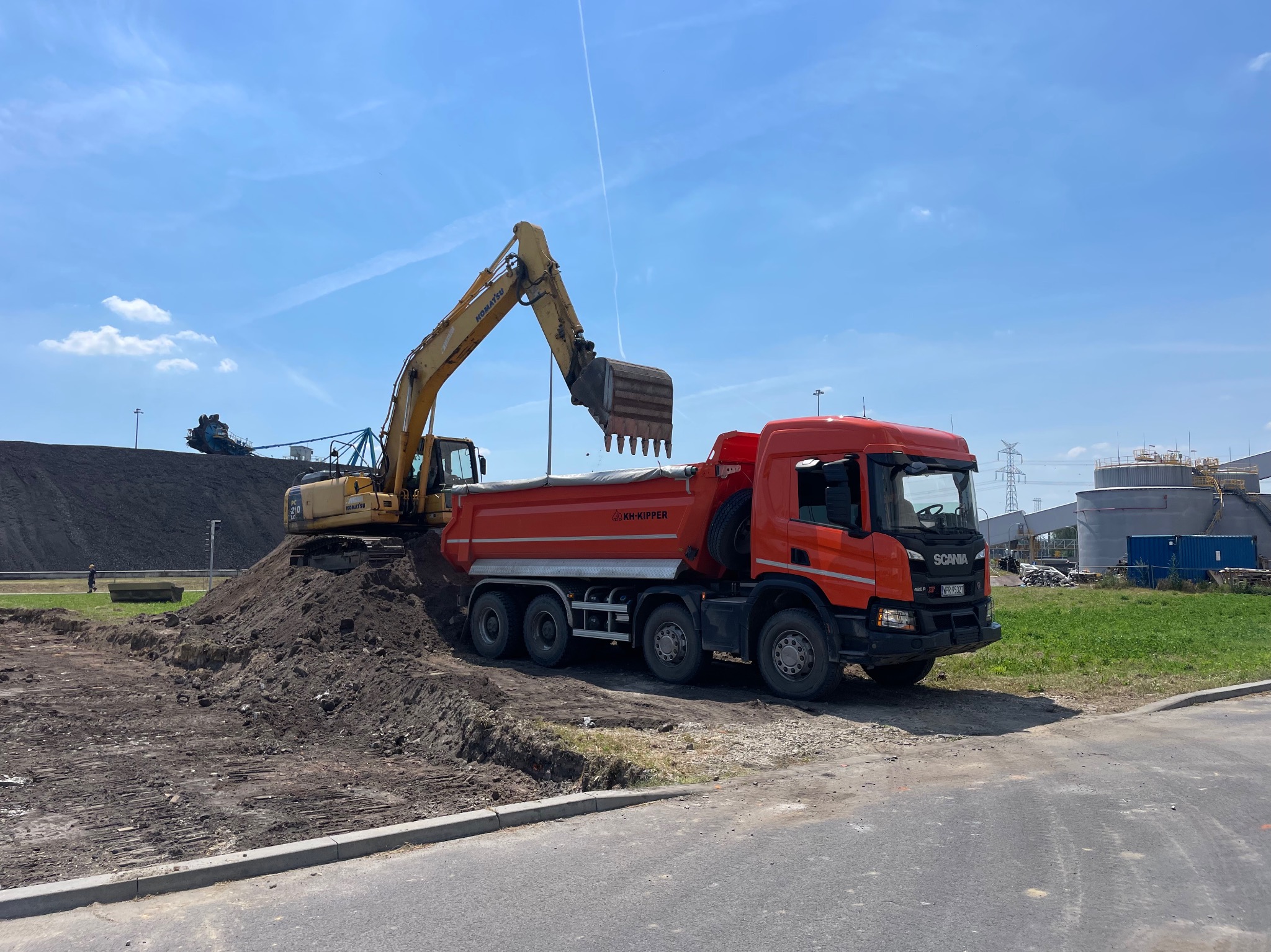 Żółta koparka Komatsu i czerwona wywrotka Scania na placu budowy w Jaworznie. Koparka nabiera ziemię z hałdy, przygotowując teren pod inwestycję.