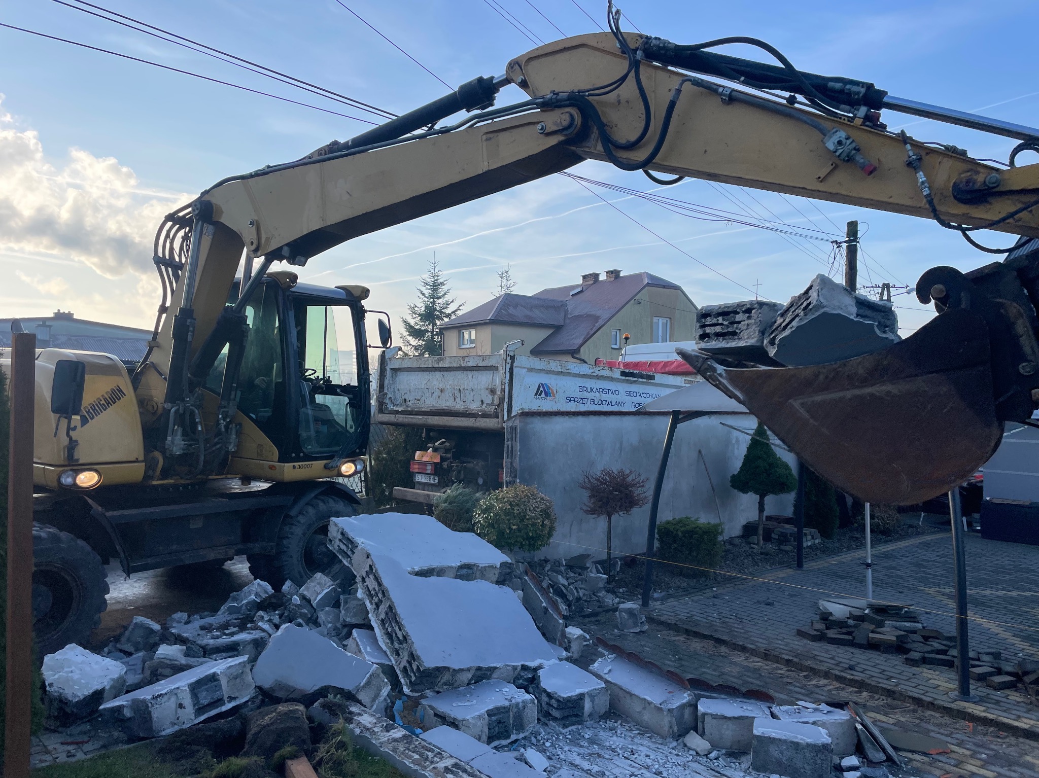Żółta koparka z łyżką nabiera gruz na tle budynku i niebieskiego nieba. Widoczne fragmenty rozbiórki i ciężarówka. Prace ziemne w toku.