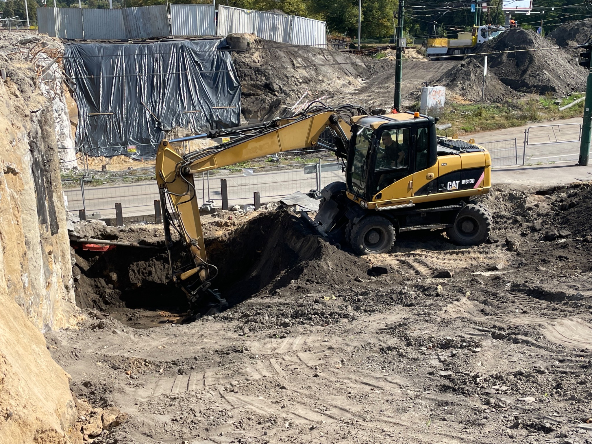 Żółta koparka CAT M313D w trakcie prac ziemnych, wykopu. W tle zabezpieczona ściana wykopu i hałda ziemi. Widoczne ślady opon na ziemi.