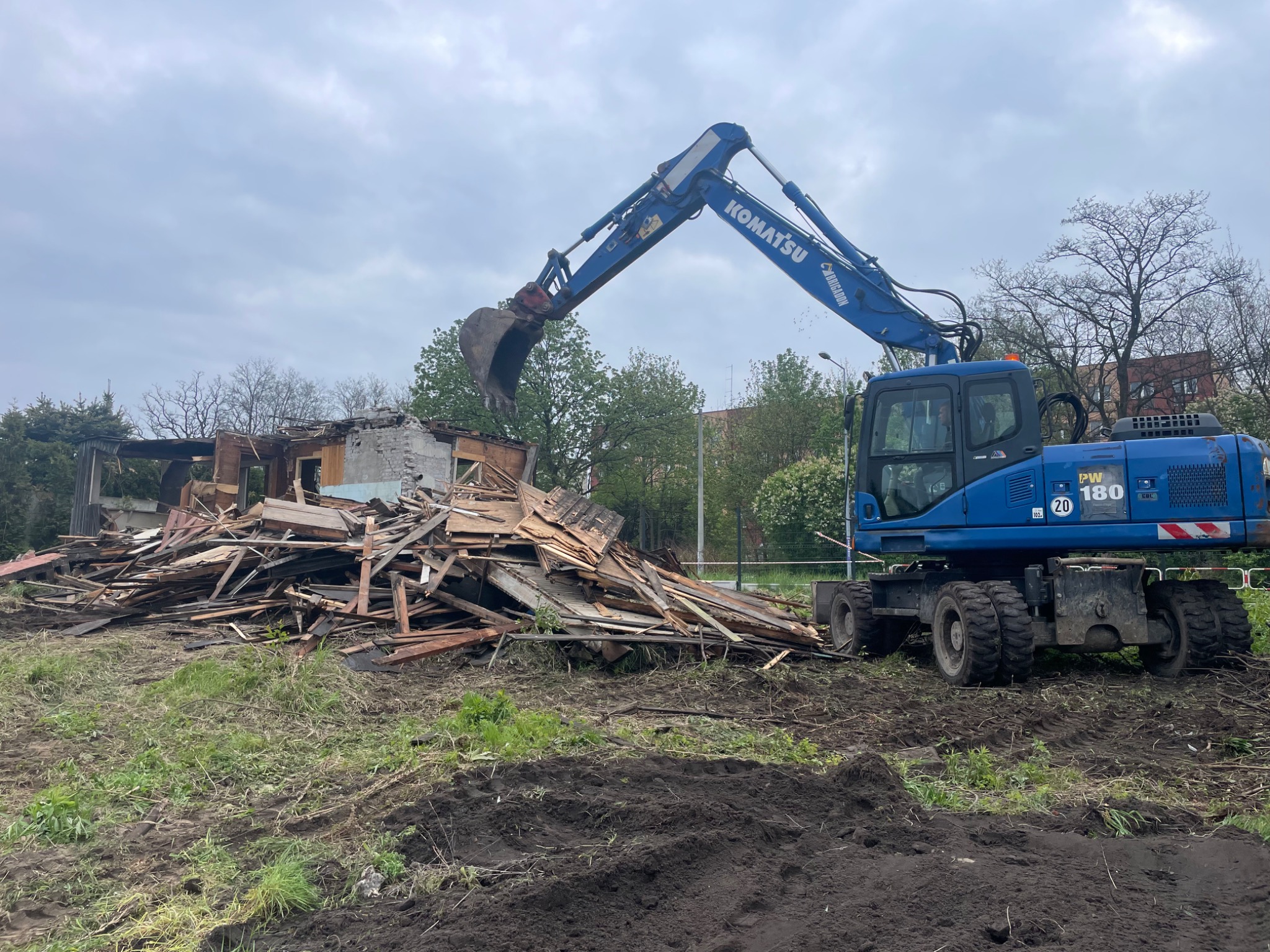 Niebieska koparka Komatsu PW180 podczas rozbiórki drewnianego budynku. Gruz i resztki konstrukcji na ziemi, pochmurne niebo w tle. Widoczne ślady opon na błotnistym terenie.