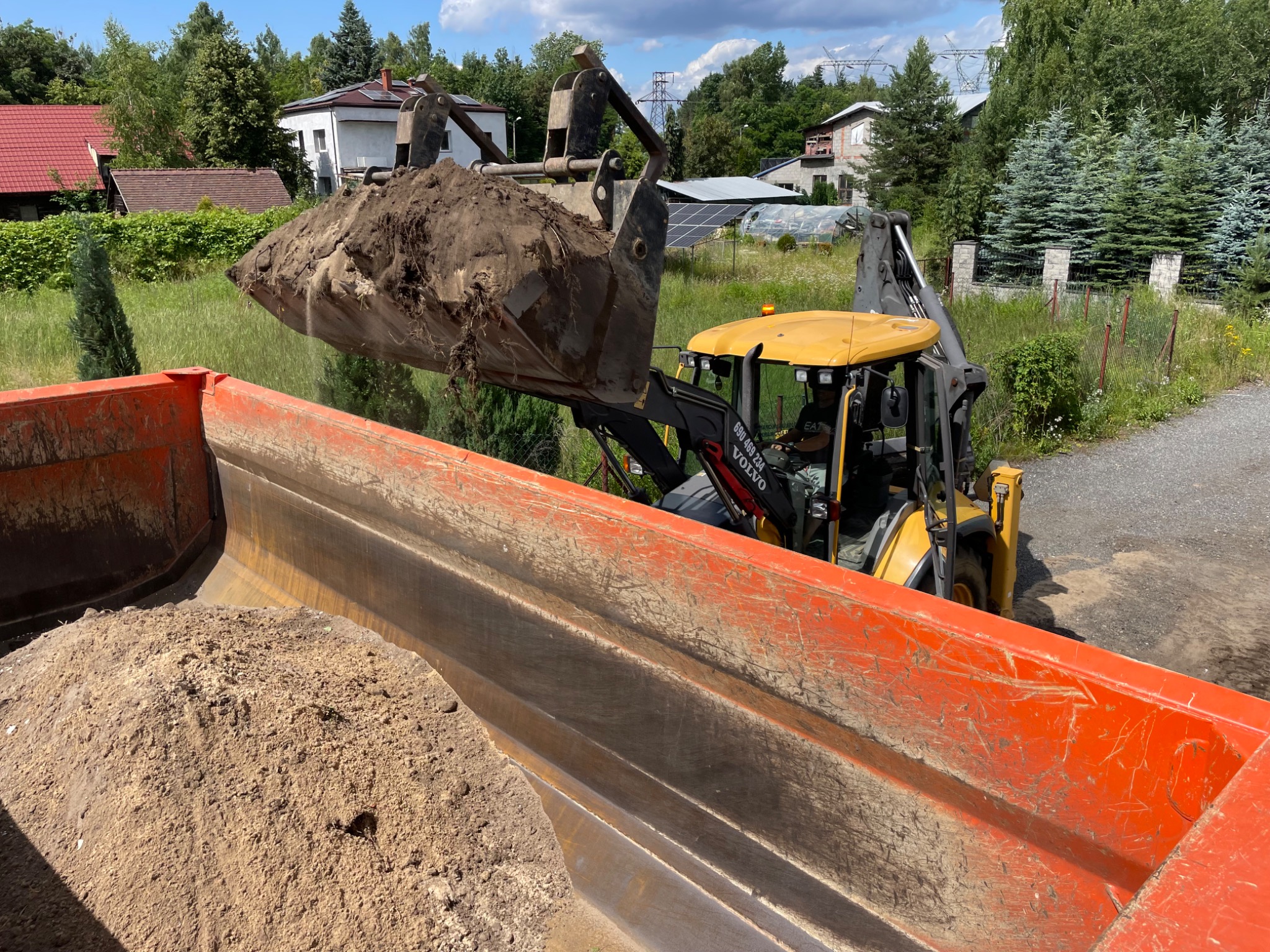 Żółta koparko-ładowarka Volvo z pełną łyżką ziemi nad pomarańczową skrzynią wywrotki, w tle zabudowa jednorodzinna i zieleń. Słoneczny dzień.