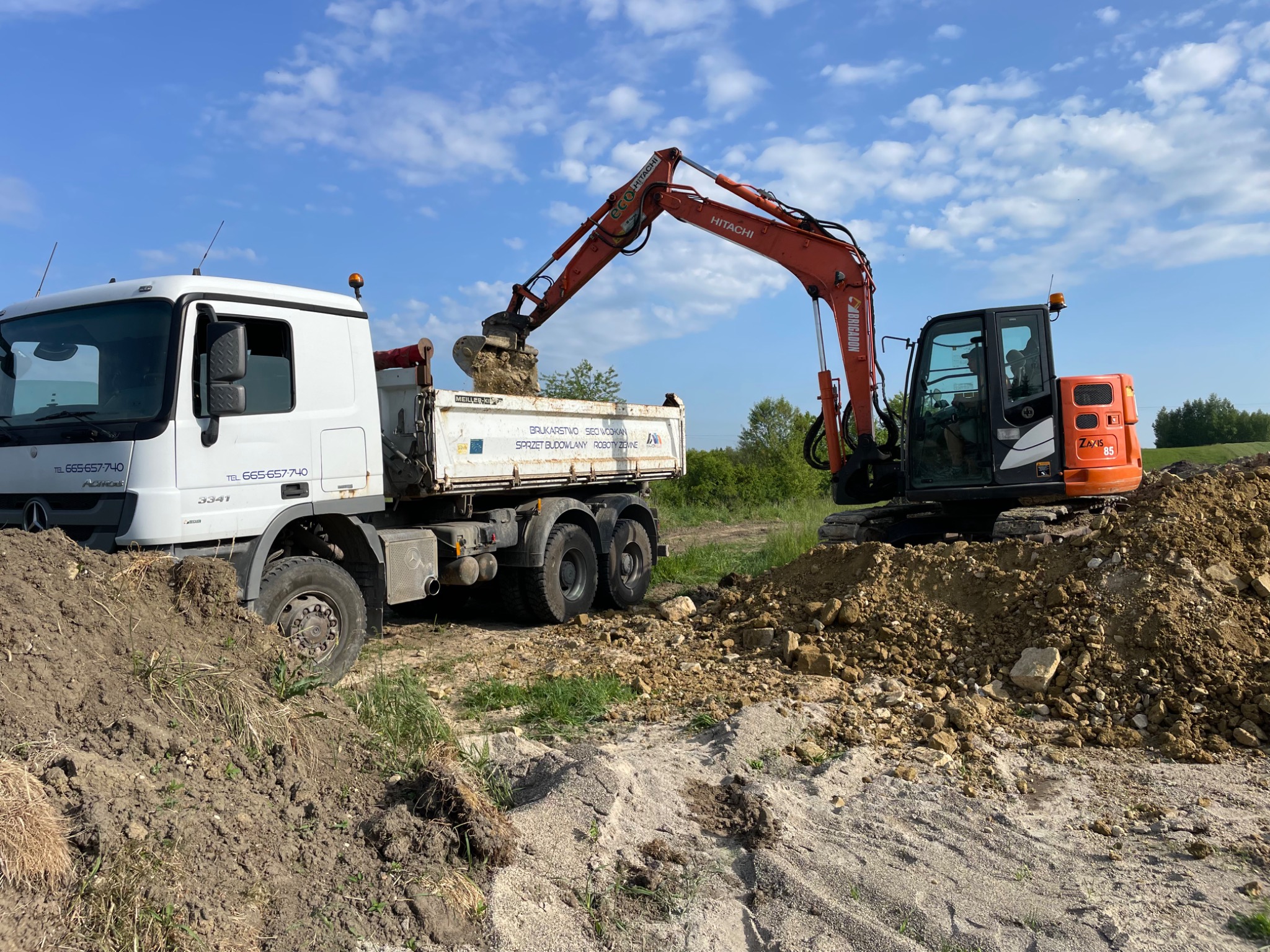 Pomarańczowa koparka Hitachi nabiera ziemię do białej ciężarówki Mercedes z niebieskimi napisami na tle błękitnego nieba. Roboty ziemne w toku.
