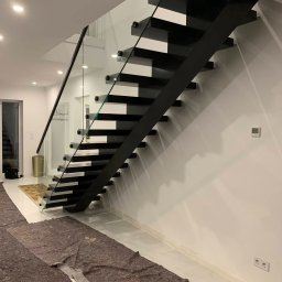 PROJEKT SCHODY Tomasz Bednarz - Nowoczesne schody wspornikowe z czarną konstrukcją i szklaną balustradą. Widoczne zabezpieczenia podłogi podczas montażu. Minimalistyczny design.