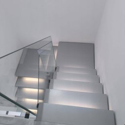 PROJEKT SCHODY Tomasz Bednarz - Nowoczesne schody z podświetleniem LED i szklaną balustradą, widok z dołu, minimalistyczny design, jasne kolory, marmurowe wykończenie podłogi.