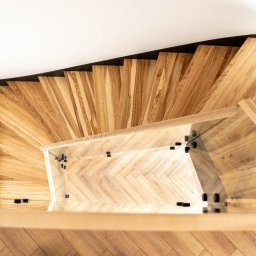 PROJEKT SCHODY Tomasz Bednarz - Drewniane schody kręcone z balustradą ze szkła, widok z góry. Stopnie ułożone w jodełkę, czarne elementy mocujące szkło. Nowoczesny design, jasne drewno.