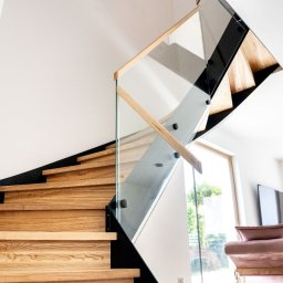 PROJEKT SCHODY Tomasz Bednarz - Nowoczesne schody z drewnianymi stopniami i szklaną balustradą, mocowaną na czarnych wspornikach. Widok z dołu, jasne wnętrze z oknami w tle.