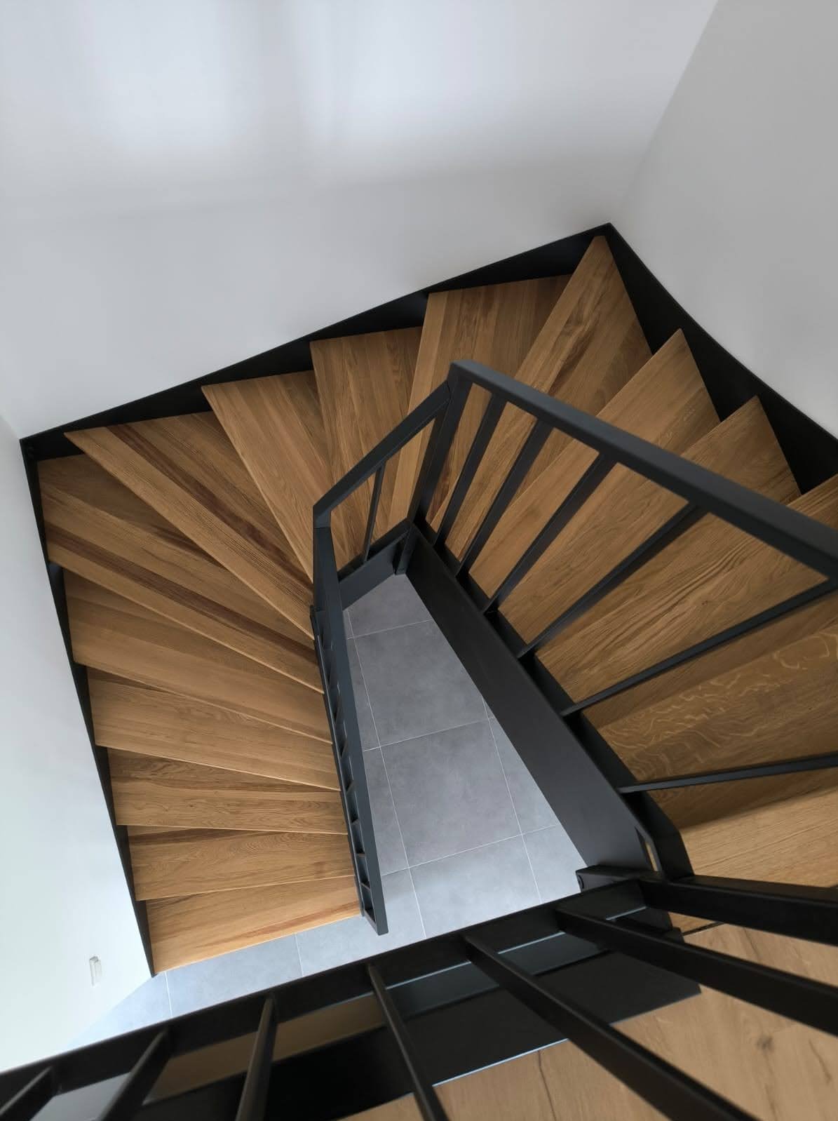 Spiralne schody z drewna dębowego z czarną, metalową balustradą, widziane z góry. Minimalistyczny design, jasne drewno, szare płytki na podeście.