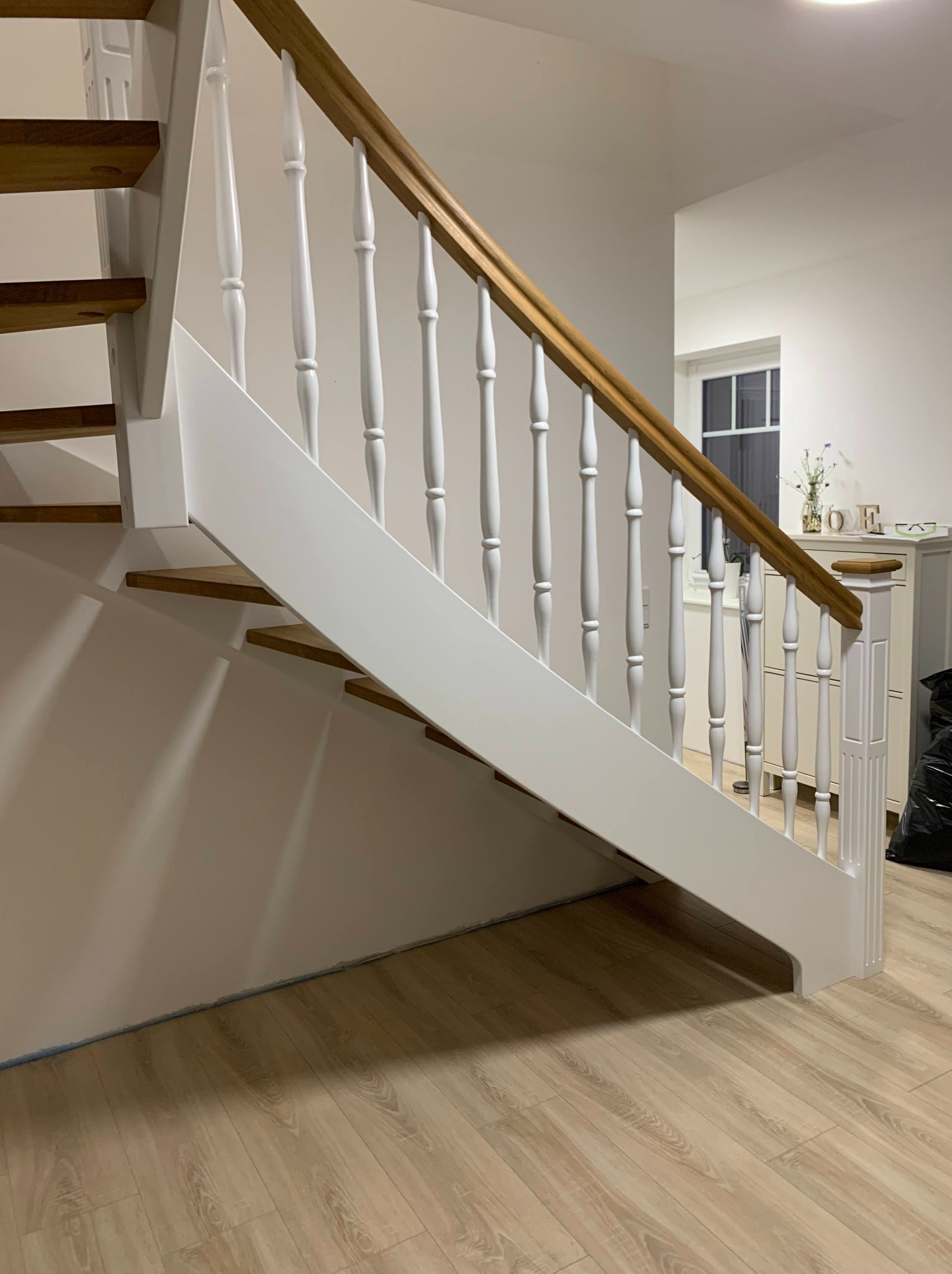 Drewniane schody z białymi balustradami i dębowym poręczą w jasnym wnętrzu. Widoczne drewniane stopnie i fragment podłogi. Minimalistyczny design.