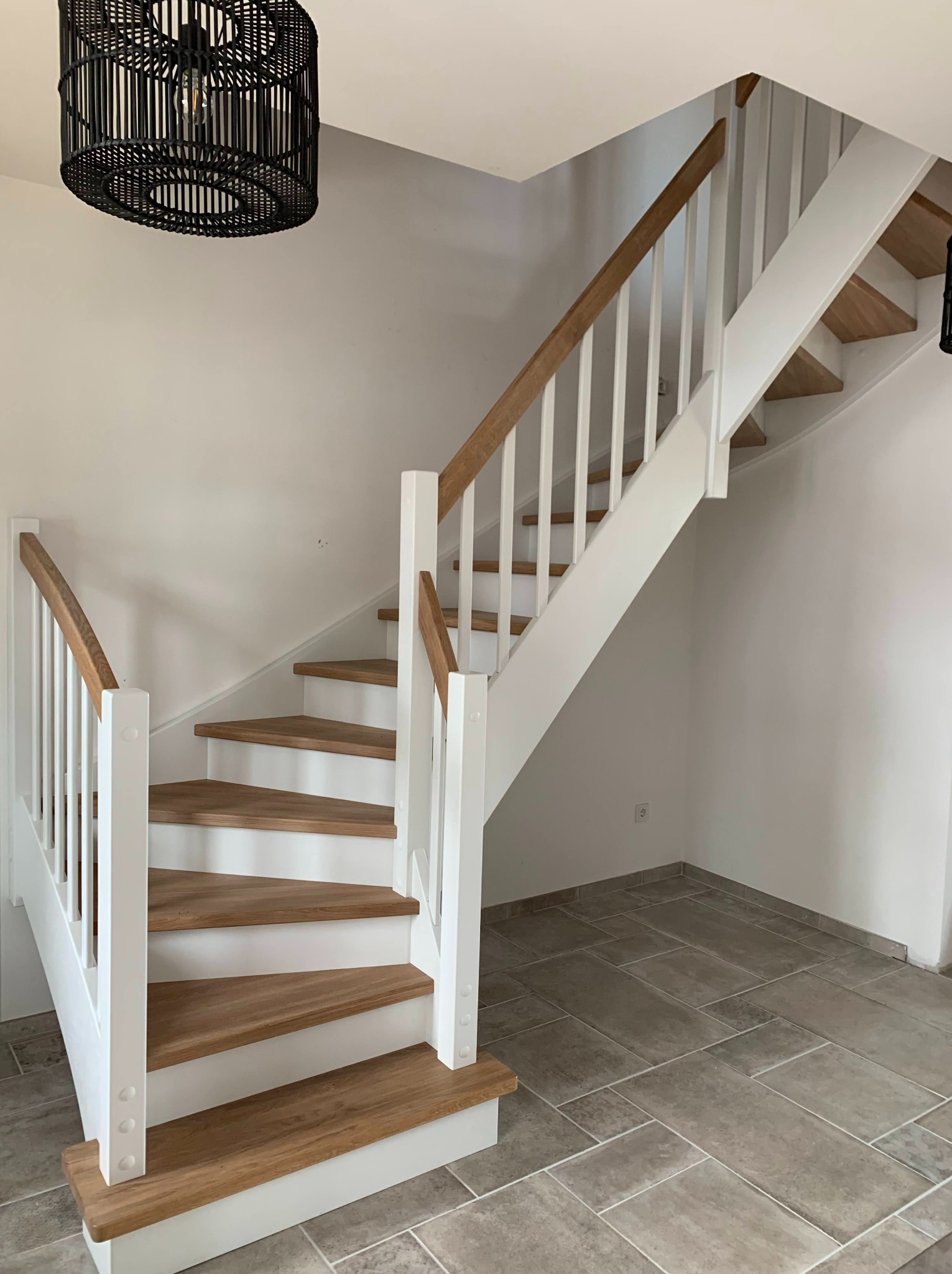 Jasne schody z drewnianymi stopniami i białą balustradą, oświetlone czarną lampą wiszącą. Widok z dołu, ukazujący detale wykończenia i minimalistyczny design.