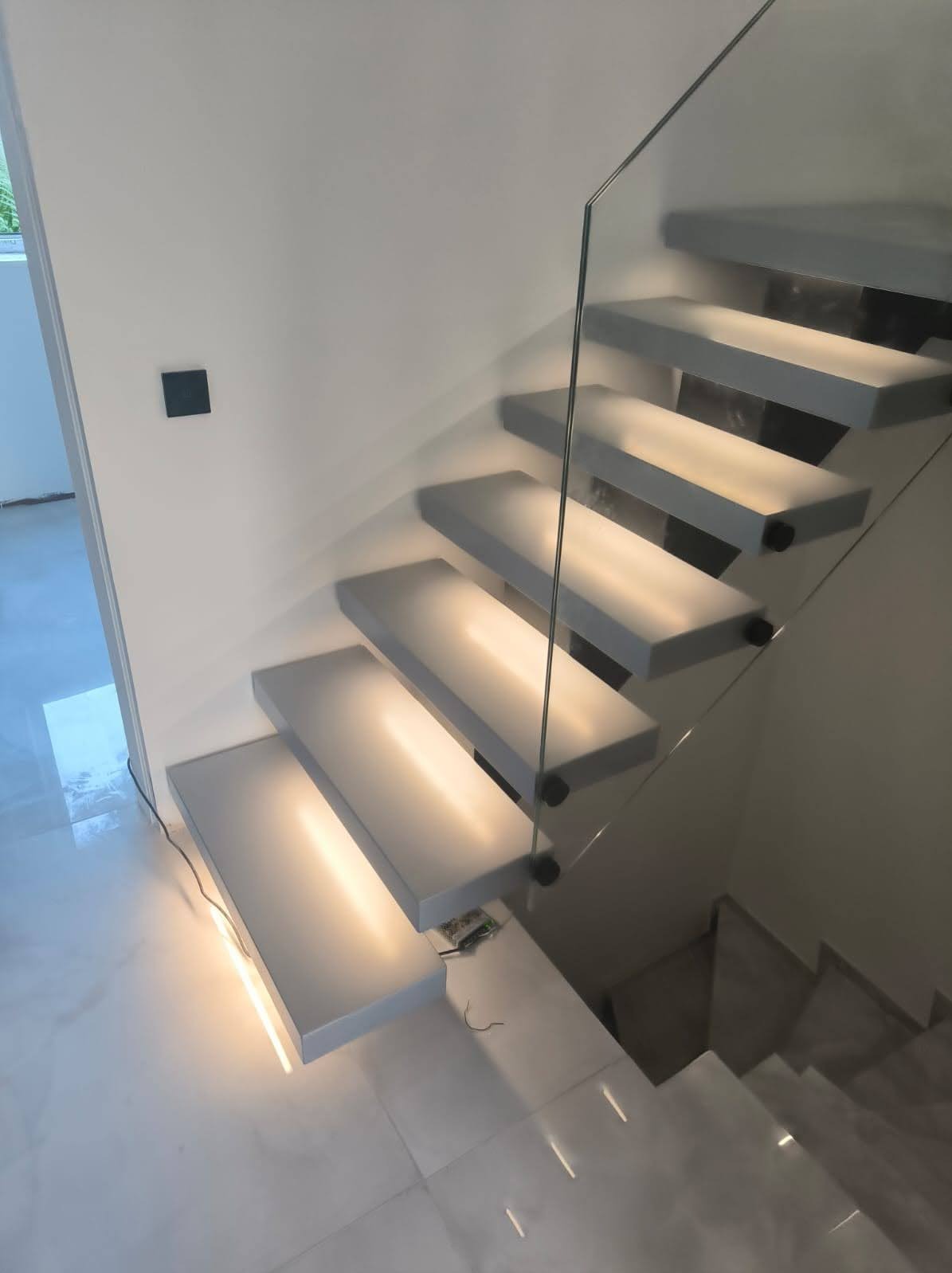 Nowoczesne schody wspornikowe z podświetleniem LED i szklaną balustradą. Minimalistyczny design, jasne wnętrze, marmurowe płytki podłogowe.