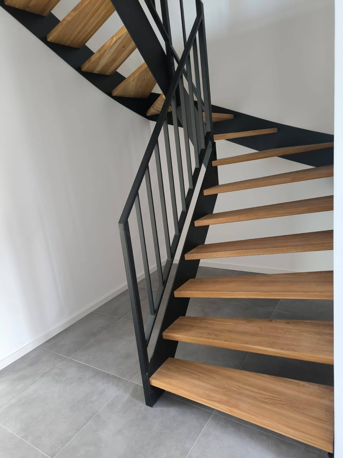 Nowoczesne schody z drewnianymi stopniami i czarną metalową balustradą, widok z dołu, szare płytki podłogowe, białe ściany, minimalistyczny design.