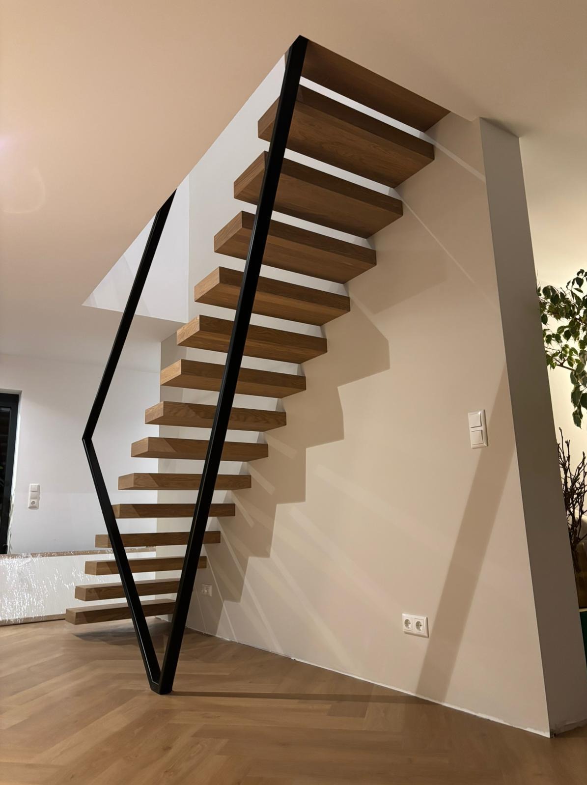 Nowoczesne schody z drewnianymi stopniami i czarną, geometryczną balustradą. Minimalistyczny design, jasne wnętrze, parkiet w jodełkę. Widok z dołu, oświetlenie naturalne.