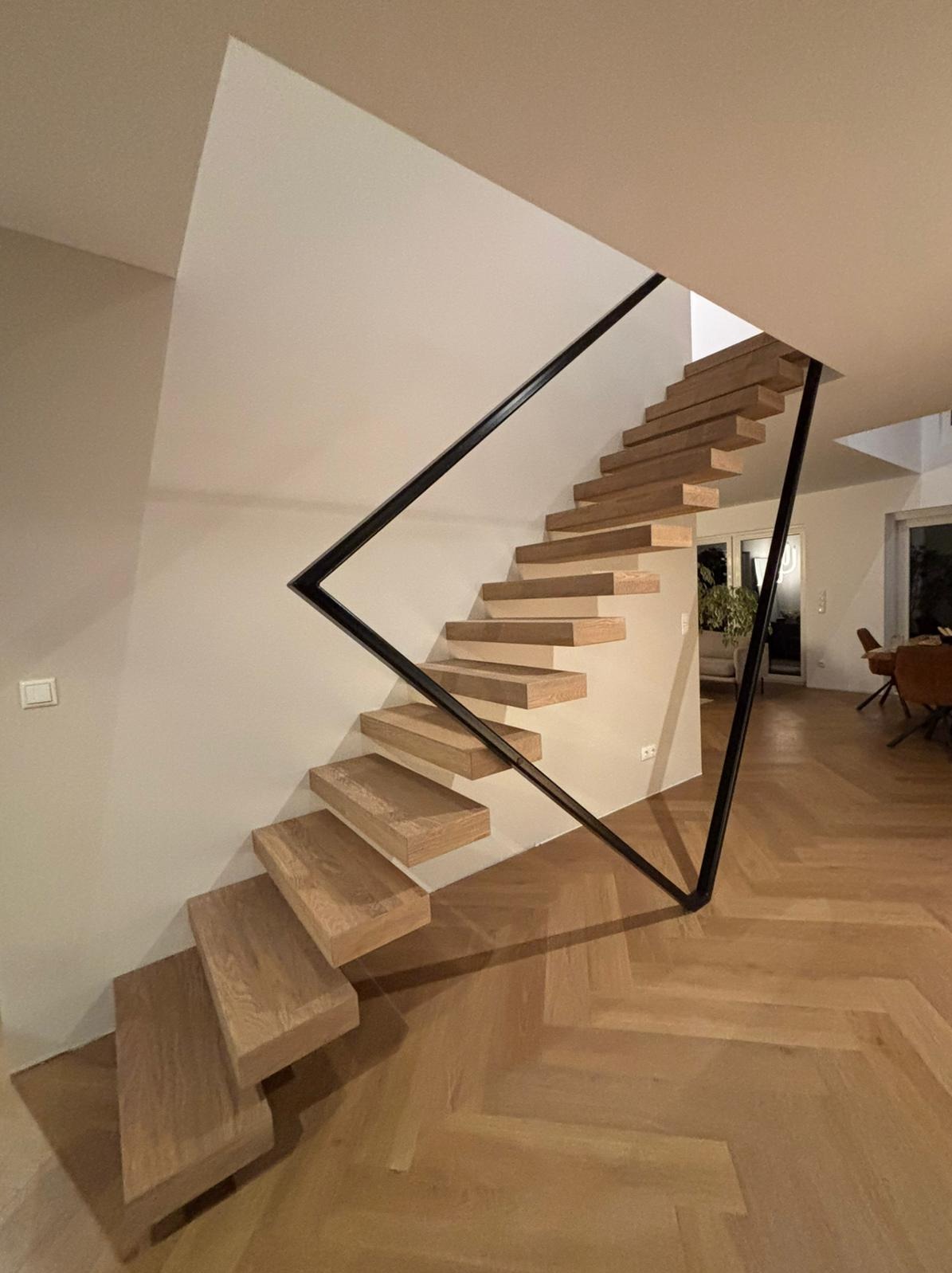 Nowoczesne schody wspornikowe z jasnego drewna i czarną, geometryczną balustradą. Parkiet w jodełkę na podłodze. Minimalistyczny design, ciepłe oświetlenie.