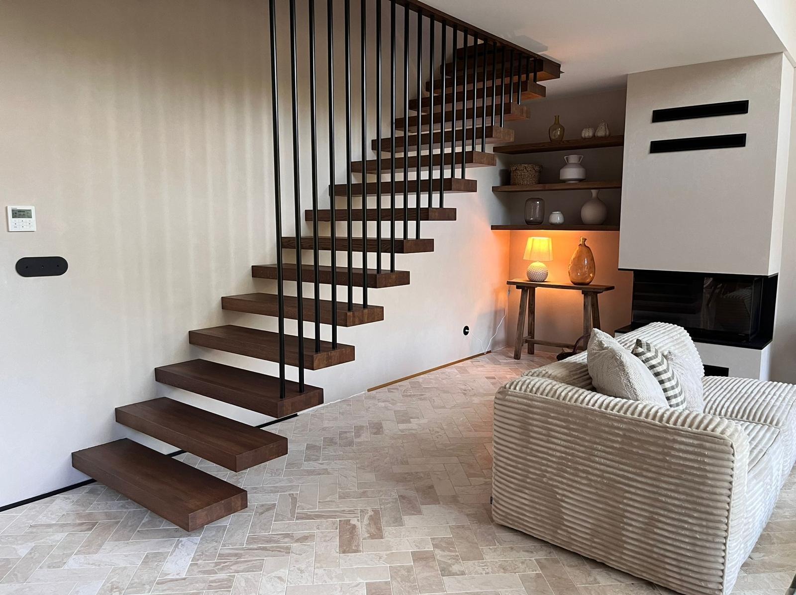 Nowoczesne schody wspornikowe z ciemnego drewna i czarną balustradą, w jasnym wnętrzu z kremową sofą i kominkiem. Minimalistyczny design, ciepłe oświetlenie.