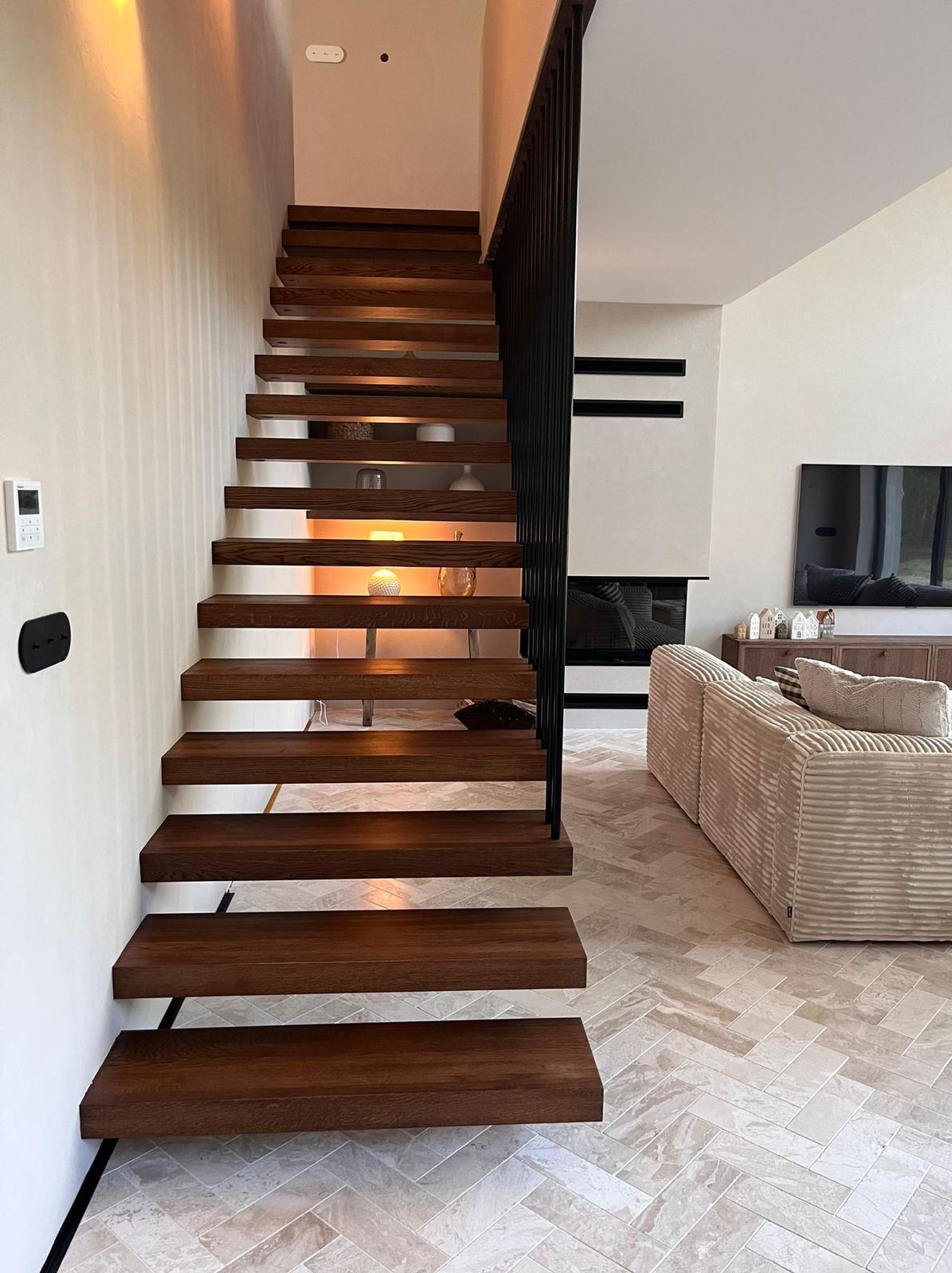 Nowoczesne schody drewniane z podświetleniem w Kołobrzegu. Stopnie z ciemnego drewna, minimalistyczna czarna balustrada. W tle salon z kominkiem i jasną podłogą.