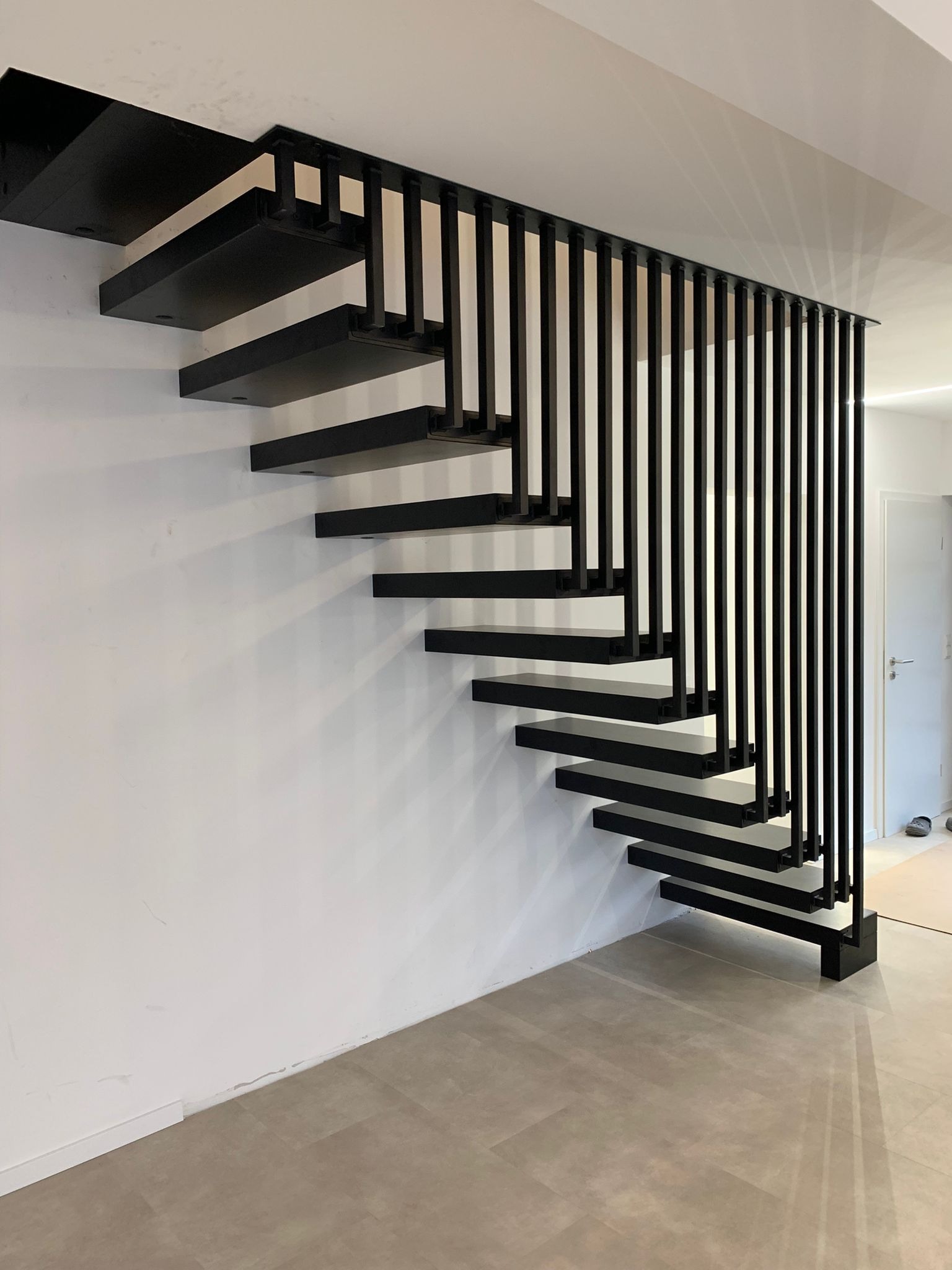 Nowoczesne, czarne schody wspornikowe z metalową balustradą z pionowych prętów, zamontowane przy białej ścianie. Minimalistyczny design, jasne wnętrze.