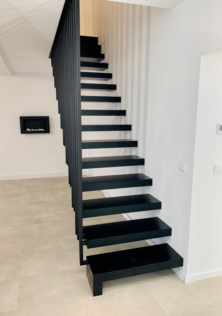 Nowoczesne, czarne schody wspornikowe z metalową balustradą w minimalistycznym wnętrzu. Widoczne subtelne pionowe pasy na ścianie i dekoracyjny kominek.