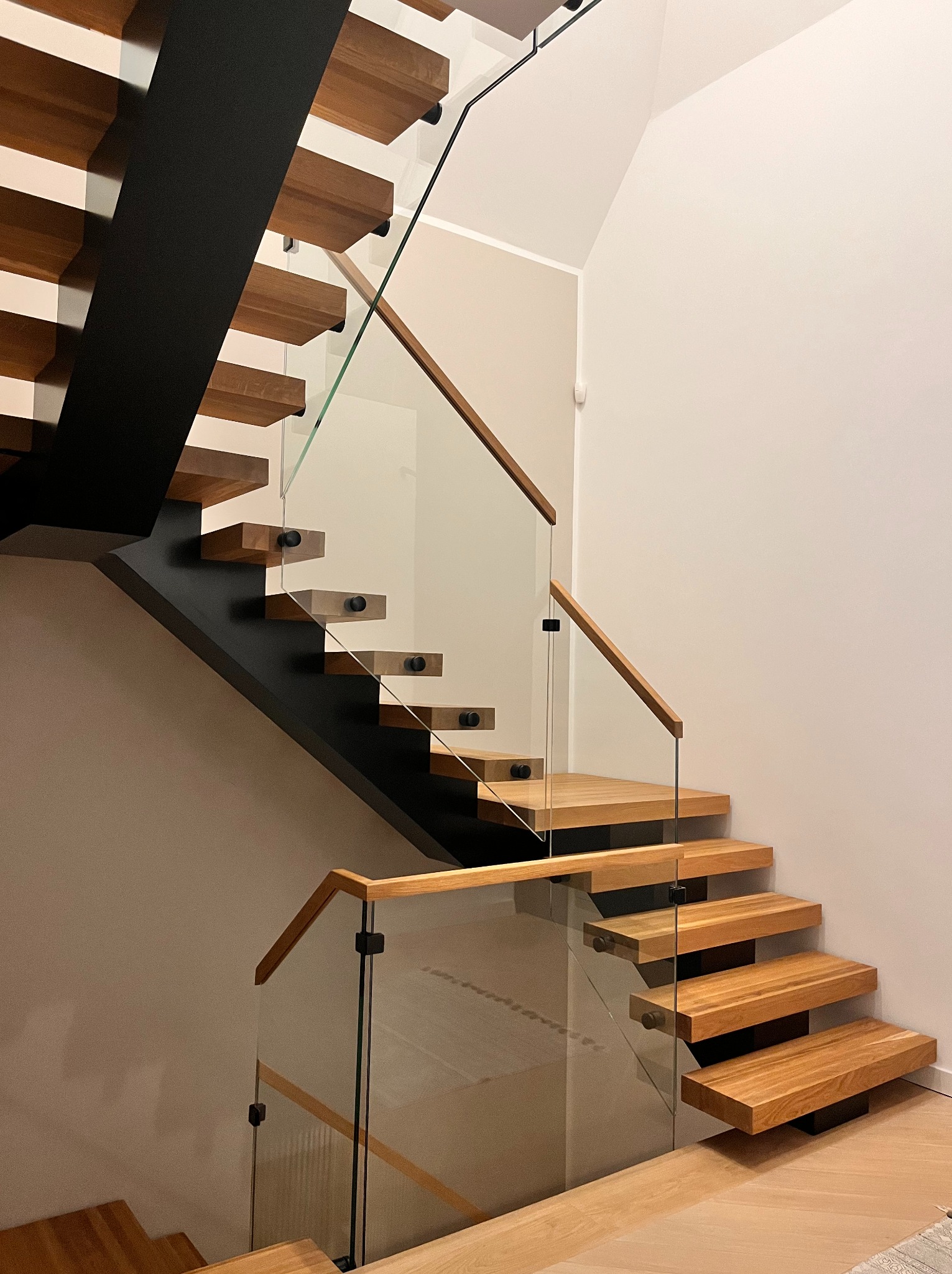 Nowoczesne schody z drewnianymi stopniami i szklaną balustradą, wsparte na czarnej, metalowej konstrukcji. Minimalistyczny design, jasne drewno, subtelne oświetlenie.