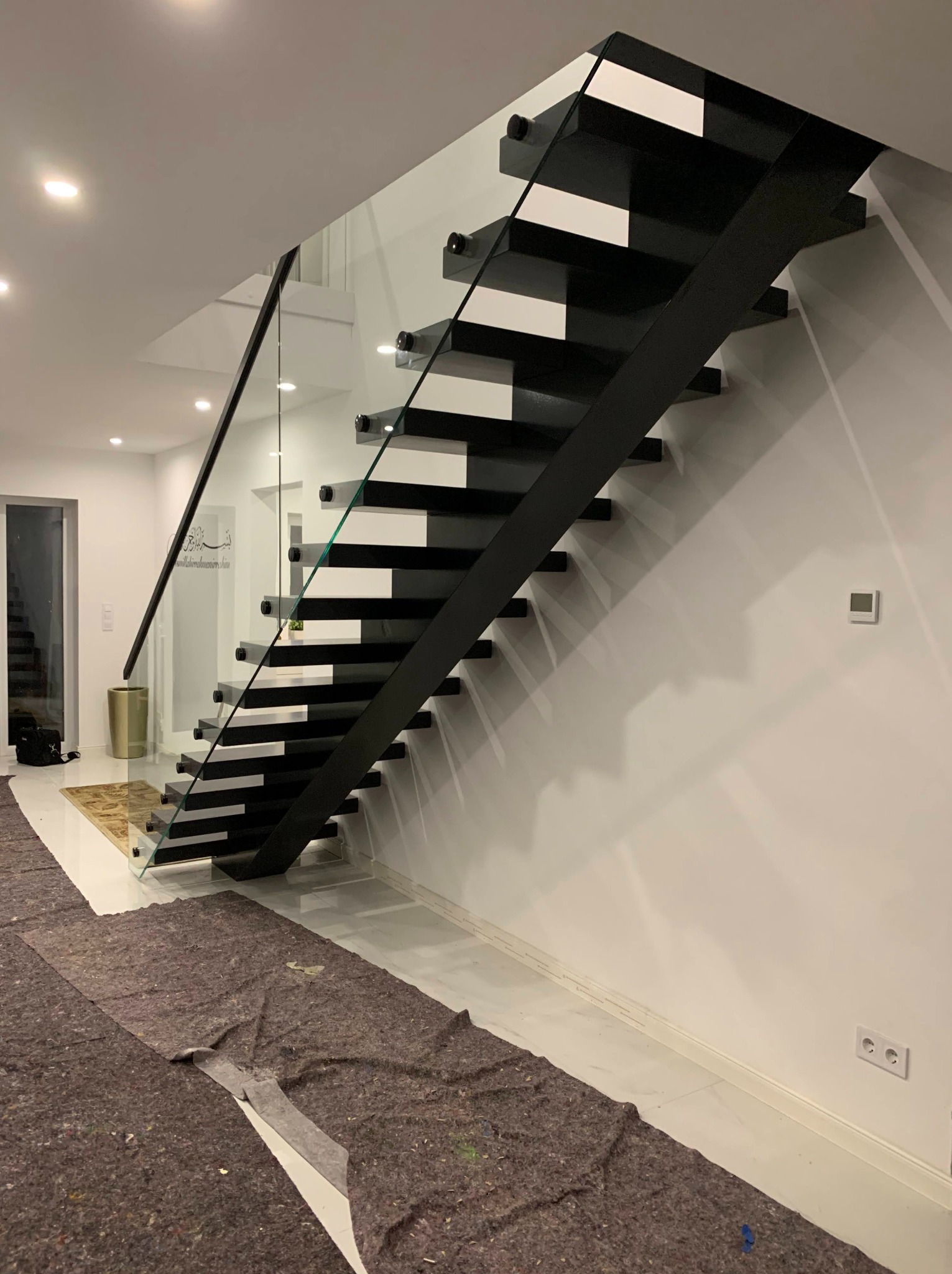 Nowoczesne schody wspornikowe z czarną konstrukcją i szklaną balustradą. Widoczne zabezpieczenia podłogi podczas montażu. Minimalistyczny design.