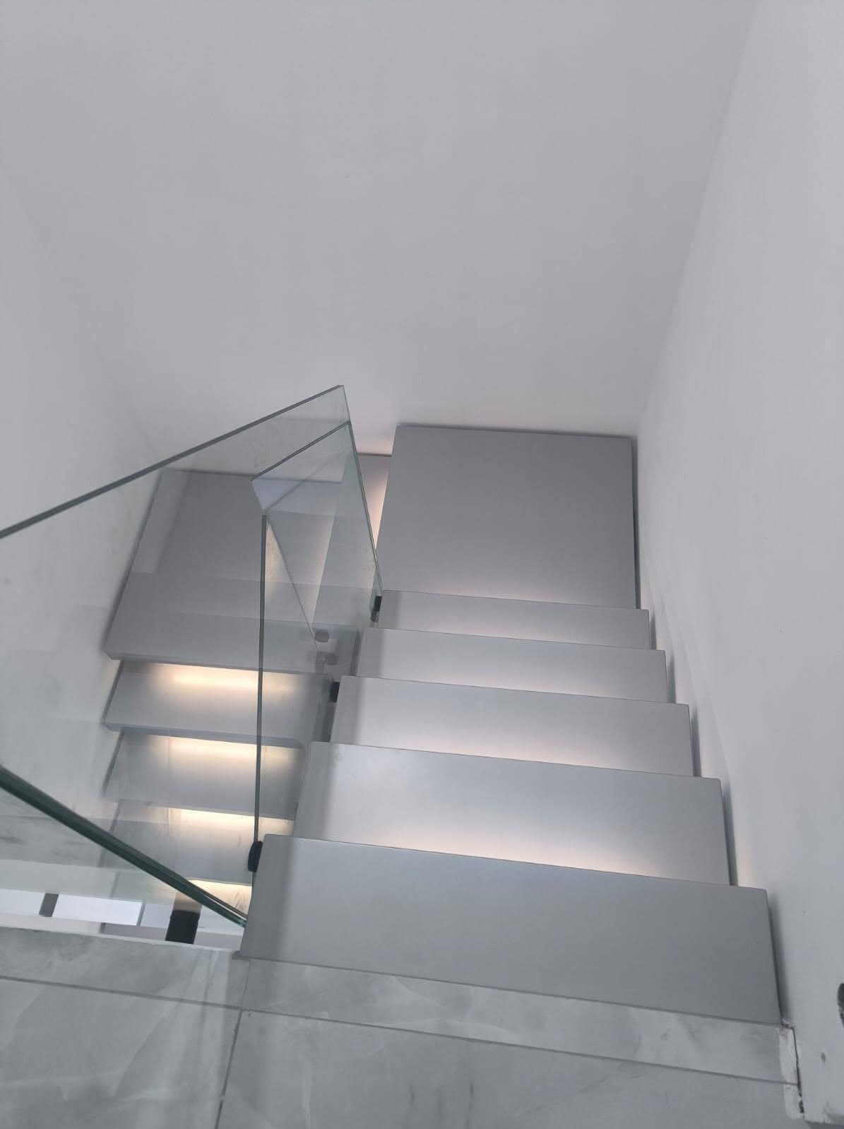 Nowoczesne schody z podświetleniem LED i szklaną balustradą, widok z dołu, minimalistyczny design, jasne kolory, marmurowe wykończenie podłogi.