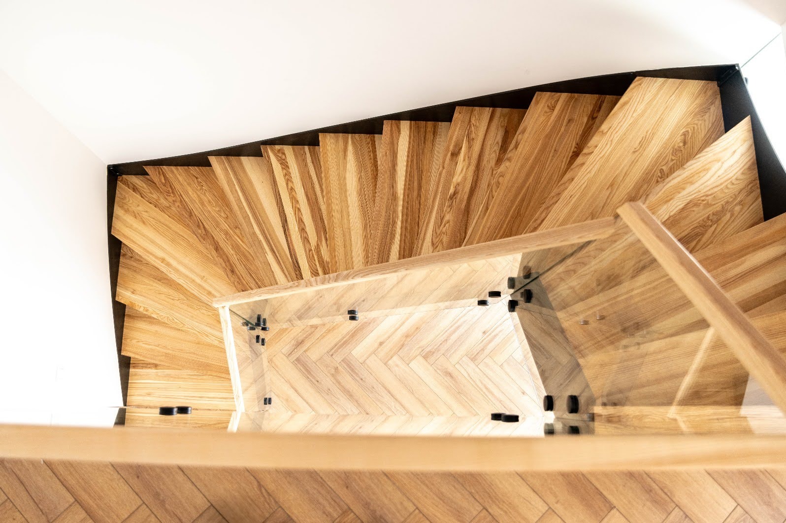 Drewniane schody kręcone z balustradą ze szkła, widok z góry. Stopnie ułożone w jodełkę, czarne elementy mocujące szkło. Nowoczesny design, jasne drewno.