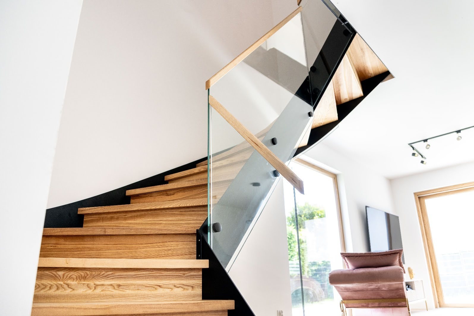 Nowoczesne schody z drewnianymi stopniami i szklaną balustradą, mocowaną na czarnych wspornikach. Widok z dołu, jasne wnętrze z oknami w tle.