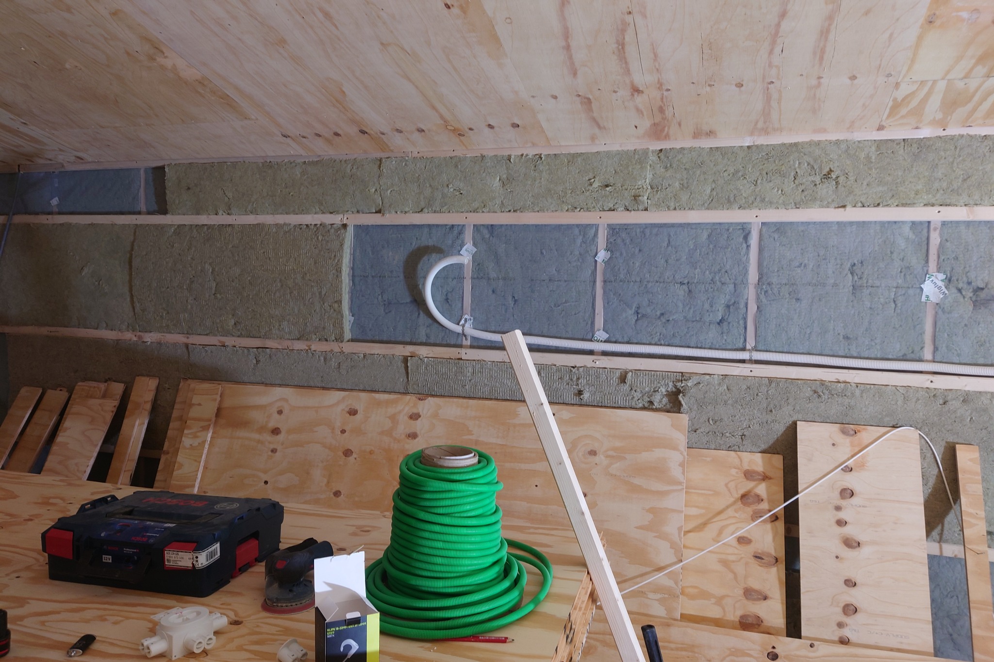 Ocieplenie poddasza wełną mineralną, widoczne przewody elektryczne, narzędzia i materiały budowlane. Prace wykończeniowe na drewnianym poddaszu.