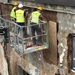 KRB POLSKA SPÓŁKA Z OGRANICZONĄ ODPOWIEDZIALNOŚCIĄ - Pracownicy na podnośniku koszowym, w żółtych kamizelkach, prowadzą prace remontowe elewacji budynku z odsłoniętą warstwą izolacyjną i fragmentem okna.