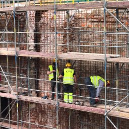 KRB POLSKA SPÓŁKA Z OGRANICZONĄ ODPOWIEDZIALNOŚCIĄ - Pracownicy w żółtych kamizelkach na rusztowaniu, demontaż ceglanej ściany z siatką wzmacniającą. Widoczne narzędzia i fragmenty usuniętej ściany.