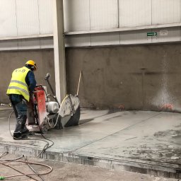 KRB POLSKA SPÓŁKA Z OGRANICZONĄ ODPOWIEDZIALNOŚCIĄ - Pracownik w żółtej kamizelce tnie betonową posadzkę przecinarką z tarczą diamentową w hali przemysłowej. Widoczne linie cięcia i rozpryskująca woda.