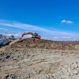 KRB POLSKA SPÓŁKA Z OGRANICZONĄ ODPOWIEDZIALNOŚCIĄ - Pomarańczowa koparka na wzgórzu ziemi, kontenery budowlane w tle, błękitne niebo z chmurami. Prace ziemne w toku, widok na budynki w oddali.