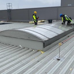 KRB POLSKA SPÓŁKA Z OGRANICZONĄ ODPOWIEDZIALNOŚCIĄ - Dwóch pracowników w uprzężach na dachu z blachy trapezowej, montujących świetlik dachowy. Widoczne zabezpieczenia linowe i narzędzia pracy.