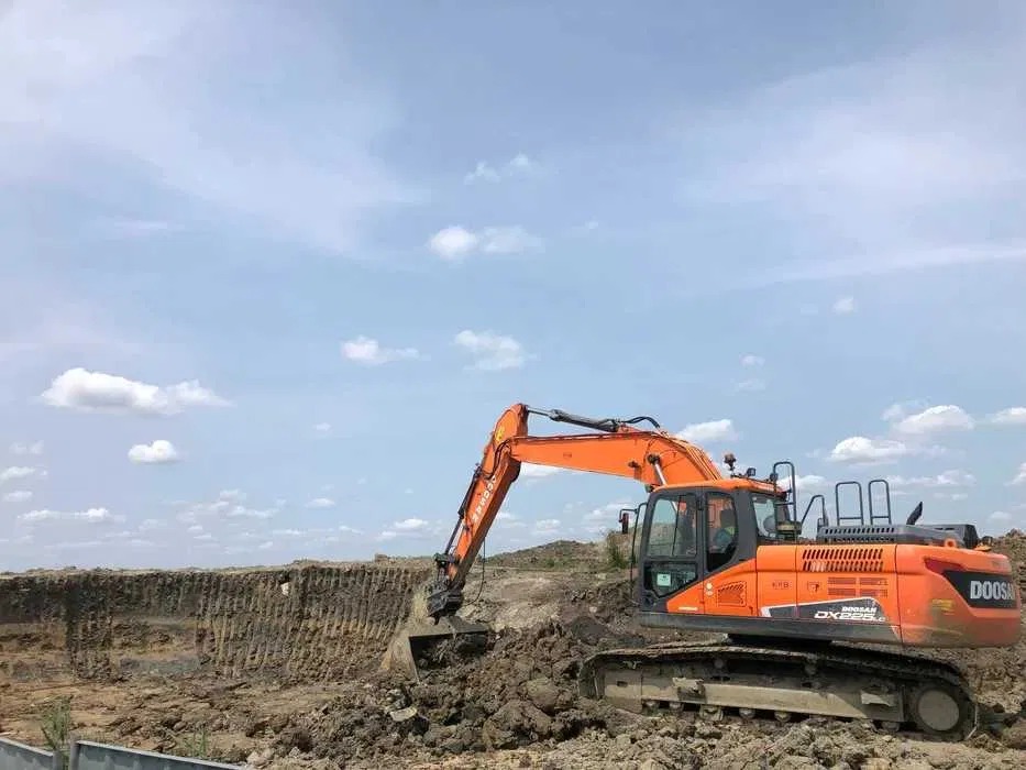 Pomarańczowa koparka Doosan DX225LC w trakcie prac ziemnych, widoczny wykop w ziemi, błękitne niebo z chmurami w tle. Praca w toku.