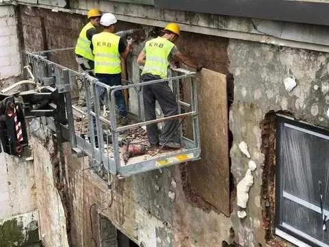 Pracownicy na podnośniku koszowym, w żółtych kamizelkach, prowadzą prace remontowe elewacji budynku z odsłoniętą warstwą izolacyjną i fragmentem okna.