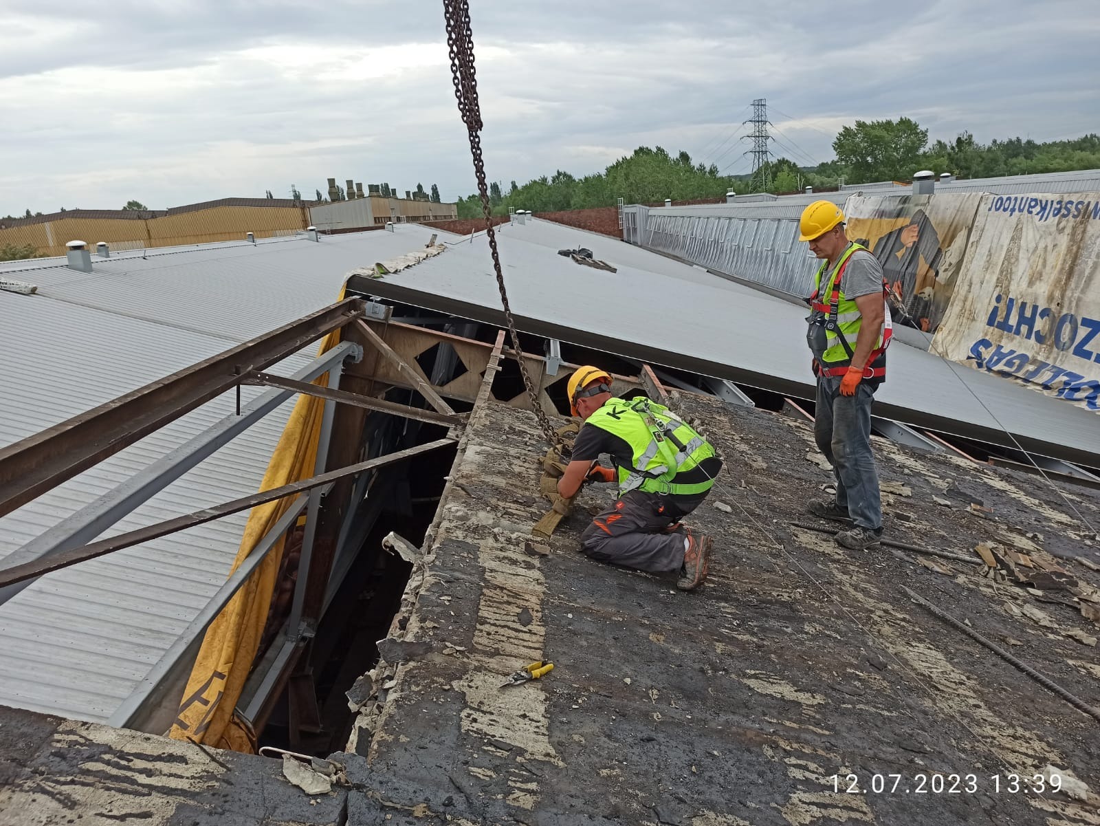 Demontaż dachu przez dwóch pracowników w kaskach i kamizelkach w Katowicach. Widoczny fragment konstrukcji i łańcuch. Prace rozbiórkowe w toku, pochmurne niebo.