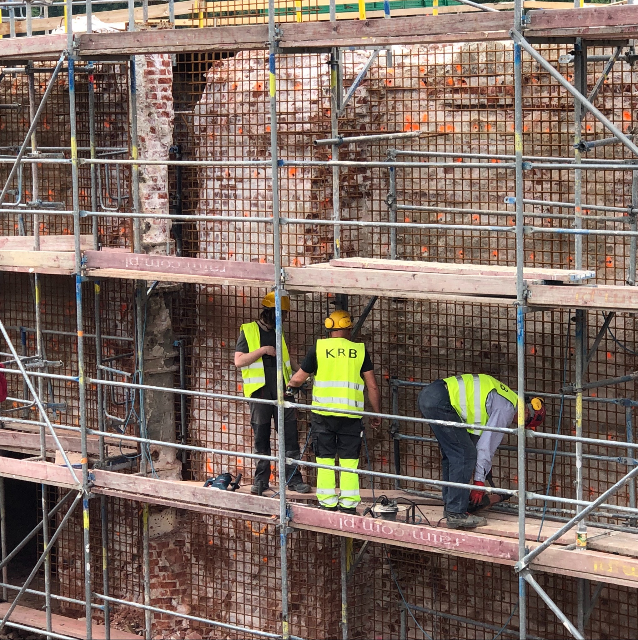 Pracownicy w żółtych kamizelkach na rusztowaniu, demontaż ceglanej ściany z siatką wzmacniającą. Widoczne narzędzia i fragmenty usuniętej ściany.